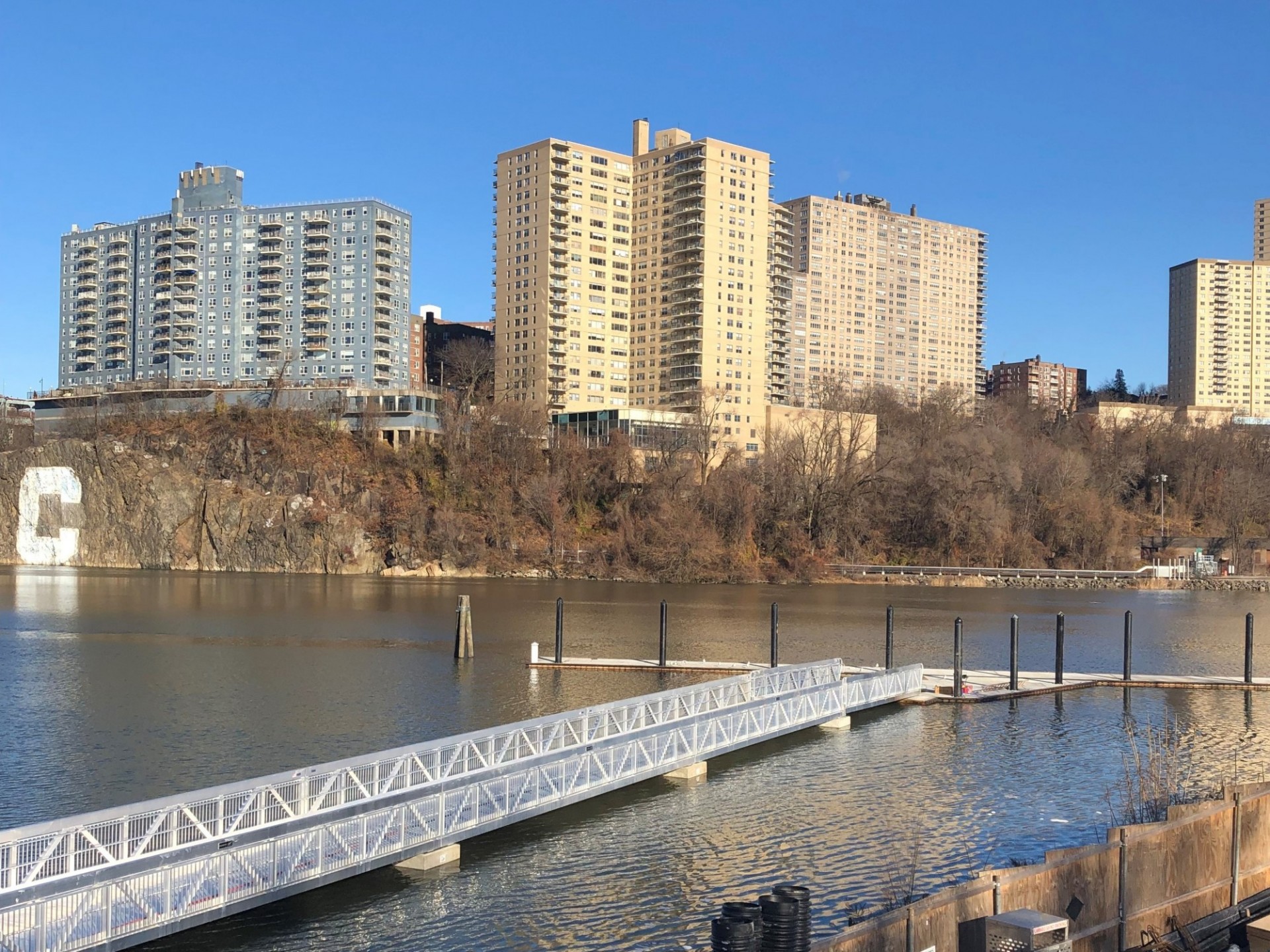 A long metal dock that extends into the Harlem River, with apartment buildings on the other side of the shore.