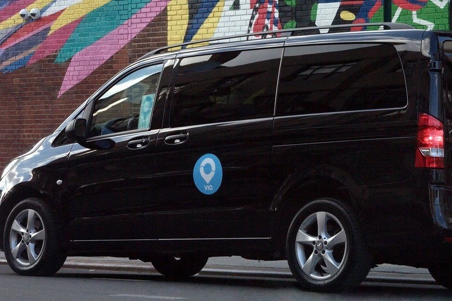 A black van that has a blue Via logo on the door is parked on a street that has a colorful street art mural on the wall behind it.