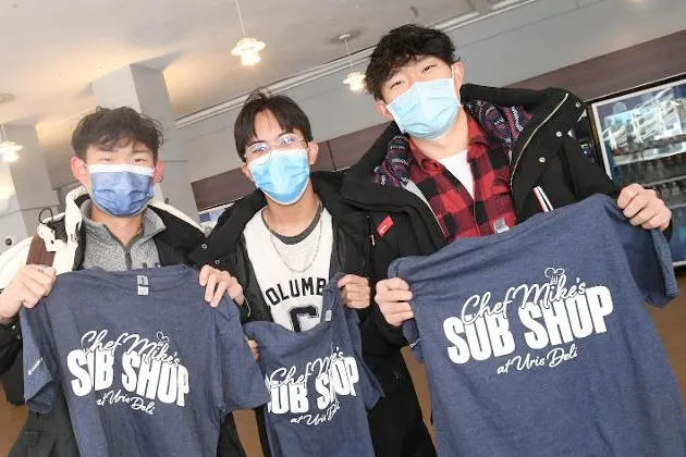 Three Columbia students gather together and hold blue shirts that say Chef Mike's Sub Shop at Uris Deli.